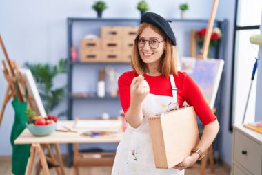 Young redhead woman at art studio holding art case beckoning come here gesture with hand inviting welcoming happy and smiling 