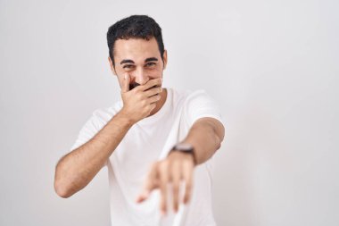Handsome hispanic man standing over white background laughing at you, pointing finger to the camera with hand over mouth, shame expression 
