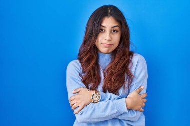 Hispanic young woman standing over blue background shaking and freezing for winter cold with sad and shock expression on face 