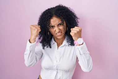 Hispanic woman with curly hair standing over pink background angry and mad raising fists frustrated and furious while shouting with anger. rage and aggressive concept. 