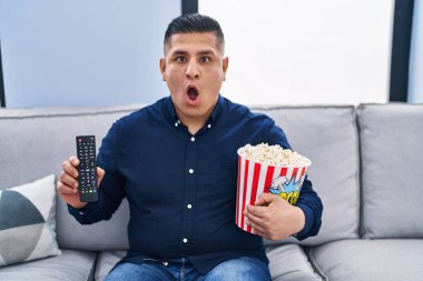 İspanyol genç adam TV kontrolü kullanarak patlamış mısır yiyor. Şaşırmış ve şaşırmış bir ifade, korku ve heyecanlı bir yüz ifadesi var.. 