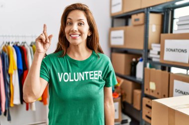 Middle age hispanic woman wearing volunteer t shirt at donations stand surprised with an idea or question pointing finger with happy face, number one 