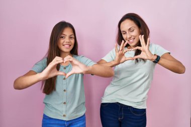 Young mother and daughter standing over pink background smiling in love doing heart symbol shape with hands. romantic concept. 