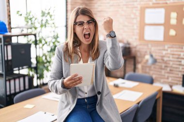 Ofiste çalışan kızgın ve kızgın gözlüklü genç İspanyol bir kadın öfkeyle bağırırken yumruğunu kaldırıyor. öfke ve agresif konsept. 