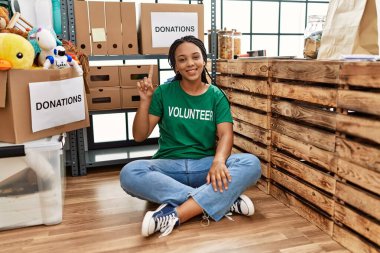 Young african american woman wearing volunteer t shirt at donations stand showing and pointing up with finger number one while smiling confident and happy. 