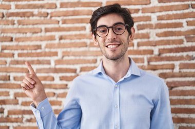 Young hispanic man standing over brick wall background with a big smile on face, pointing with hand finger to the side looking at the camera. 