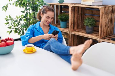 Genç bir kadın kahve içiyor ve evde akıllı telefon kullanıyor.