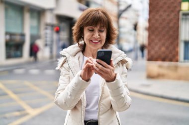 Sokakta akıllı telefon kullanan orta yaşlı bir kadın gülümsüyor.