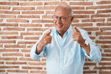 Duvarın üzerinde duran gri saçlı yaşlı adam mutlu ve komik yüzlü parmaklarını kameraya doğrultuyor. iyi enerji ve titreşimler. 
