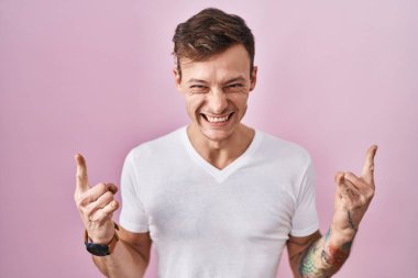 Caucasian man standing over pink background shouting with crazy expression doing rock symbol with hands up. music star. heavy music concept. 