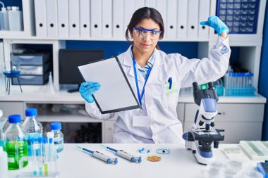 Hispanic young woman working at scientist laboratory strong person showing arm muscle, confident and proud of power 