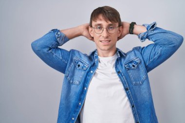 Caucasian blond man standing wearing glasses relaxing and stretching, arms and hands behind head and neck smiling happy 