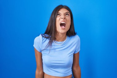 Brunette young woman standing over blue background angry and mad screaming frustrated and furious, shouting with anger. rage and aggressive concept. 