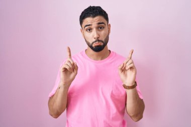 İspanyol genç adam pembe arka planda dikiliyor üzgün ve üzgün görünüyor, parmaklarıyla yön gösteriyor, mutsuz ve depresif.. 