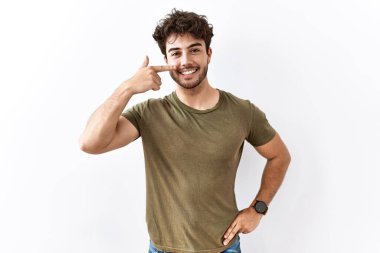 İzole edilmiş beyaz arka planda duran İspanyol bir adam, parmakları burnuna ve yüzüne dönük, neşeli bir şekilde gülümsüyor. güzellik konsepti 