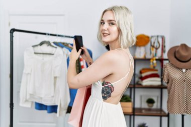 Genç beyaz kadın kendine gülerek elbise mağazasında fotoğraf çekiyor.
