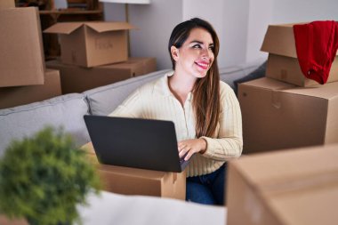 Young beautiful hispanic woman using laptop sitting on sofa at new home
