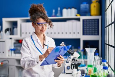 Young hispanic woman scientist smiling confident writing on document at laboratory
