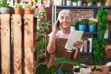 Orta yaşlı, kır saçlı, çiçekçi kadın çiçekçi çiçekçide telefon okuyup kitap okuyor.