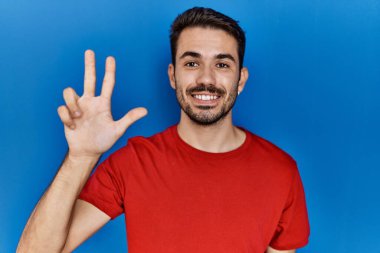 Young hispanic man with beard wearing red t shirt over blue background showing and pointing up with fingers number three while smiling confident and happy. 