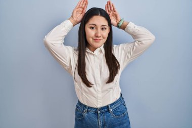 Young latin woman standing over blue background doing bunny ears gesture with hands palms looking cynical and skeptical. easter rabbit concept. 