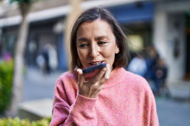 Orta yaşlı bir kadın gülümsüyor. Sokakta akıllı telefondan konuşuyor.