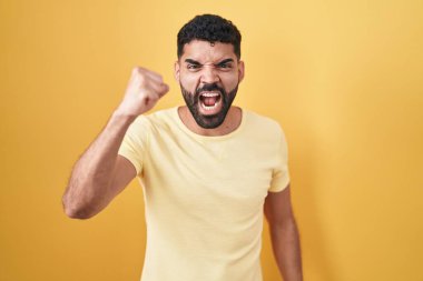Hispanic man with beard standing over yellow background angry and mad raising fist frustrated and furious while shouting with anger. rage and aggressive concept. 