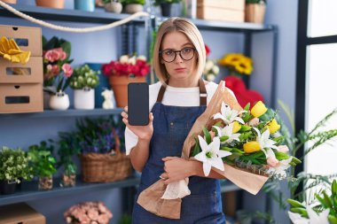 Young caucasian woman working at florist shop showing smartphone screen skeptic and nervous, frowning upset because of problem. negative person. 