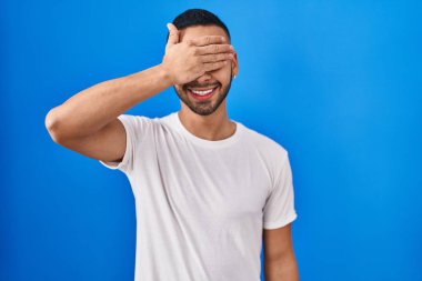 Mavi arka planda dikilen genç İspanyol adam. Gülümsüyor ve gülümsüyor. Gözlerini şaşkınlıkla kapatıyor. kör kavram. 