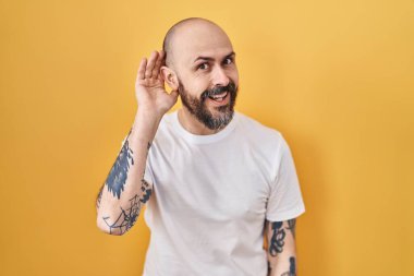 Sarı arka planda dikilen dövmeleri olan genç İspanyol bir adam. Kulağı kulağında söylentiler ve dedikoduları dinliyor. Sağırlık kavramı. 