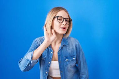 Young caucasian woman standing over blue background smiling with hand over ear listening an hearing to rumor or gossip. deafness concept. 