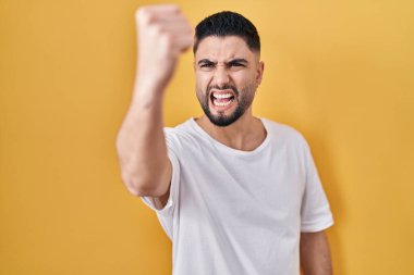 Sarı arka plan üzerine gündelik tişört giyen genç yakışıklı sinirli ve öfkeli yumruğunu kaldırırken öfkeyle bağırıyordu. öfke ve agresif konsept. 