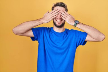 Hispanic man with beard standing over yellow background covering eyes with hands smiling cheerful and funny. blind concept. 