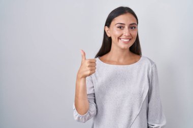 Young hispanic woman standing over white background doing happy thumbs up gesture with hand. approving expression looking at the camera showing success. 