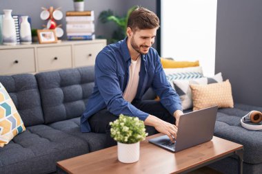 Evdeki kanepede oturan genç beyaz adam dizüstü bilgisayar kullanıyor.