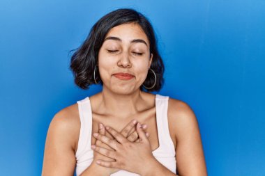 Mavi arka planda duran genç İspanyol kadın elleri göğsünde, gözleri kapalı ve yüzünde minnet dolu bir jest ile gülümsüyor. sağlık kavramı. 