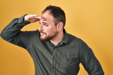 Plus size hispanic man with beard standing over yellow background very happy and smiling looking far away with hand over head. searching concept. 