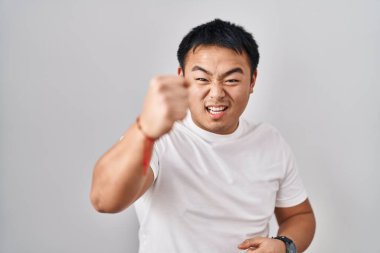 Young chinese man standing over white background angry and mad raising fist frustrated and furious while shouting with anger. rage and aggressive concept. 