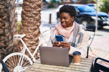 Laptop ve akıllı telefon kullanan Afro-Amerikalı bir kadın kahve dükkanının terasında oturuyor.