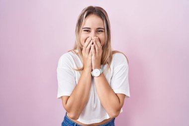 Young blonde woman standing over pink background laughing and embarrassed giggle covering mouth with hands, gossip and scandal concept 