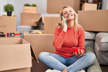 Young blonde woman talking on smartphone drinking coffee at new home