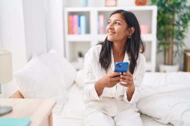 Genç, güzel, İspanyol bir kadın yatakta akıllı telefon kullanıyor.