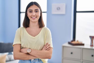 Genç İspanyol kadın gülümsüyor. Kollarını kavuşturup evde duruyor.