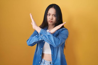 Sarı arka planda duran genç Asyalı kadın. Reddedilme ifadesi. Kollarını kavuşturma, negatif işaret, kızgın surat. 