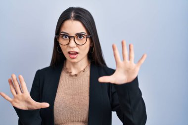 Mavi arka planda duran esmer kadın korku ifadesinden korkmuş ve dehşete düşmüş. El hareketlerini durdur, şok içinde bağır. panik konsepti. 