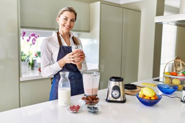 Genç sarışın kadın gülümsüyor. Mutfakta buzlu içecek bardağı tutuyor.