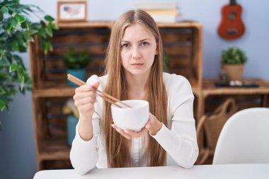 Young caucasian woman eating asian food using chopsticks skeptic and nervous, frowning upset because of problem. negative person. 