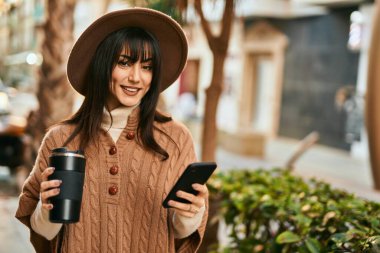 Kışlık şapkalı esmer kadın akıllı telefon kullanıyor ve şehirde bir fincan kahve içiyor.