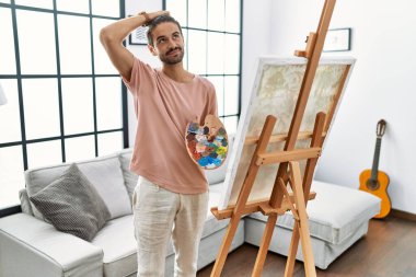 Young hispanic man with beard painting on canvas at home confuse and wondering about question. uncertain with doubt, thinking with hand on head. pensive concept. 