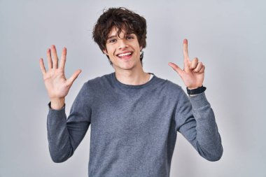 İzole edilmiş arka planda duran genç adam yedinci parmağı gösterip işaret ederken kendinden emin ve mutlu bir şekilde gülümsüyor.. 
